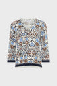 A womens V-neck top with three-quarter sleeves, featuring an intricate blue, brown, and white floral geometric pattern, and dark trim on the cuffs and hem.