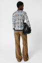 Person standing with back to camera, wearing a patterned long-sleeve top, tan flared pants, and holding a black fringed bag against a plain white background.