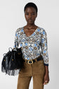 Woman in a floral V-neck shirt and brown suede pants holds a black fringed bag. She has a neutral expression and wears a black belt with a gold buckle and a gold ring, standing against a plain white background.