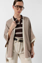 Person wearing large glasses, a brown and white striped polo shirt, a beige open cardigan layered over a stylish poncho cachemire, and high-waisted light-colored jeans with a black belt, standing against a plain white background.