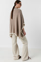 A woman with long brown hair in a ponytail wears glasses, a poncho beige with white trim—a stylish example of a poncho bicolore—paired with cream wide-leg pants and dark shoes, standing against a white background and looking to her left.