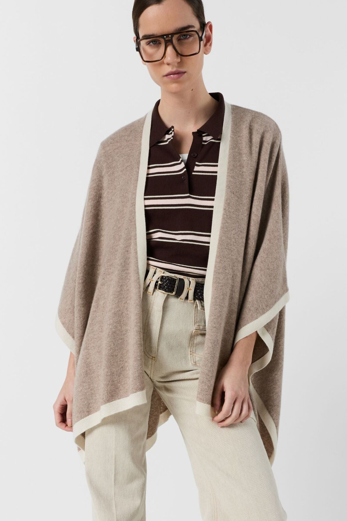 A person wearing glasses, a beige poncho cachemire over a brown and white striped shirt, and light-colored pants stands against a plain background.
