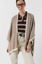 A person wearing glasses, a beige poncho cachemire over a brown and white striped shirt, and light-colored pants stands against a plain background.