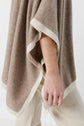 A person wearing a loose, poncho beige with white trim over light-colored pants, shown from the shoulders down with their arm relaxed at their side.