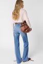 A woman with long blonde hair, wearing a light sweater and blue jeans, stands facing away, holding a large brown leather handbag. Her stylish pull col V femme adds a touch of elegance to her casual ensemble.