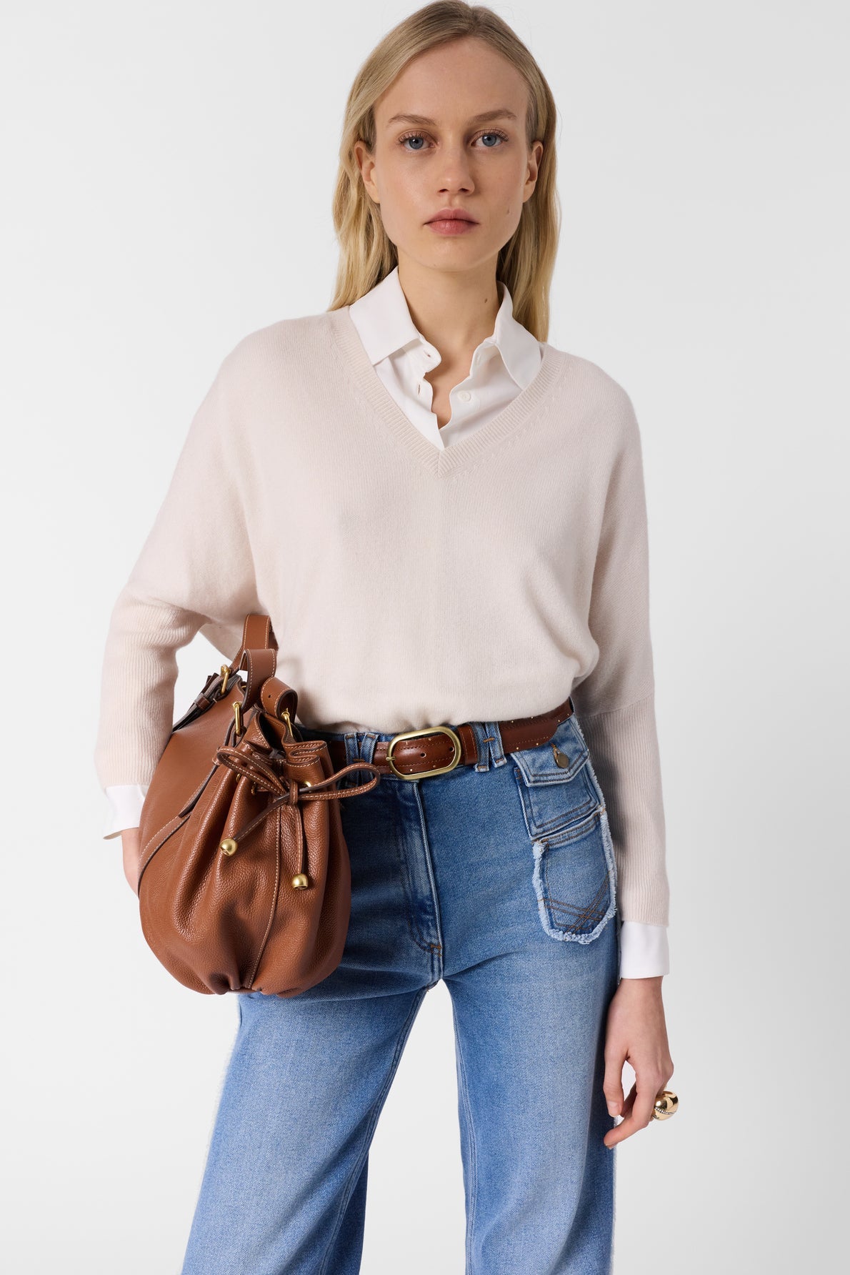 A woman wearing a light pink pull cachemire over a white shirt, blue high-waisted jeans, and a brown belt, holding a brown leather bag over her shoulder, stands against a plain white background.