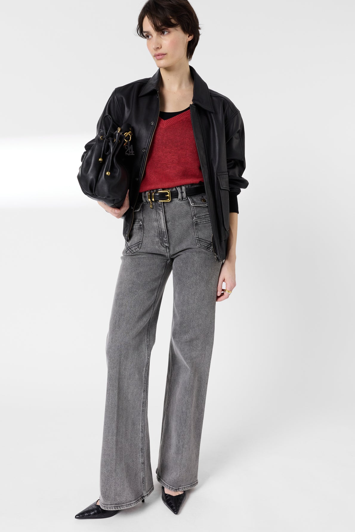 A woman wearing a black jacket, red top, gray high-waisted jeans, and black pointed shoes holds a black handbag. She stands against a plain white background.