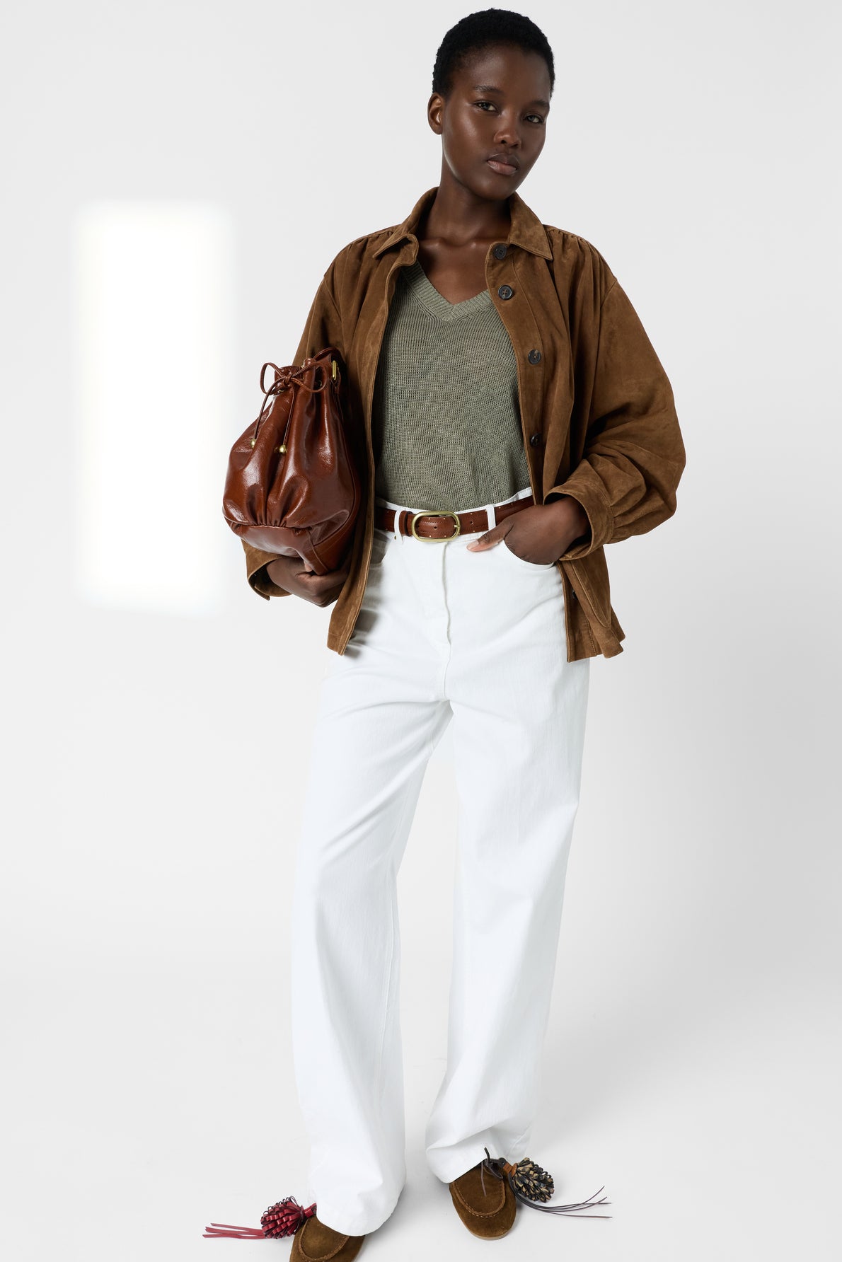 A person stands wearing a brown jacket over a pull lin femme col V kaki top, paired with white pants, a brown belt, and brown shoes, holding a brown leather bag against a plain white background.