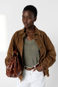 A woman in a brown suede jacket, pull lin femme with a col V in pull kaki, and white pants holds a brown leather handbag. She stands confidently against a light gray background.