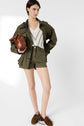 Woman stands in a khaki jacket, white V-neck top, green patterned shorts, and brown shoes. She holds a brown handbag under her arm and looks to the side against a plain white background, her outfit effortlessly complemented by a chic pull rayé femme draped over her shoulders.
