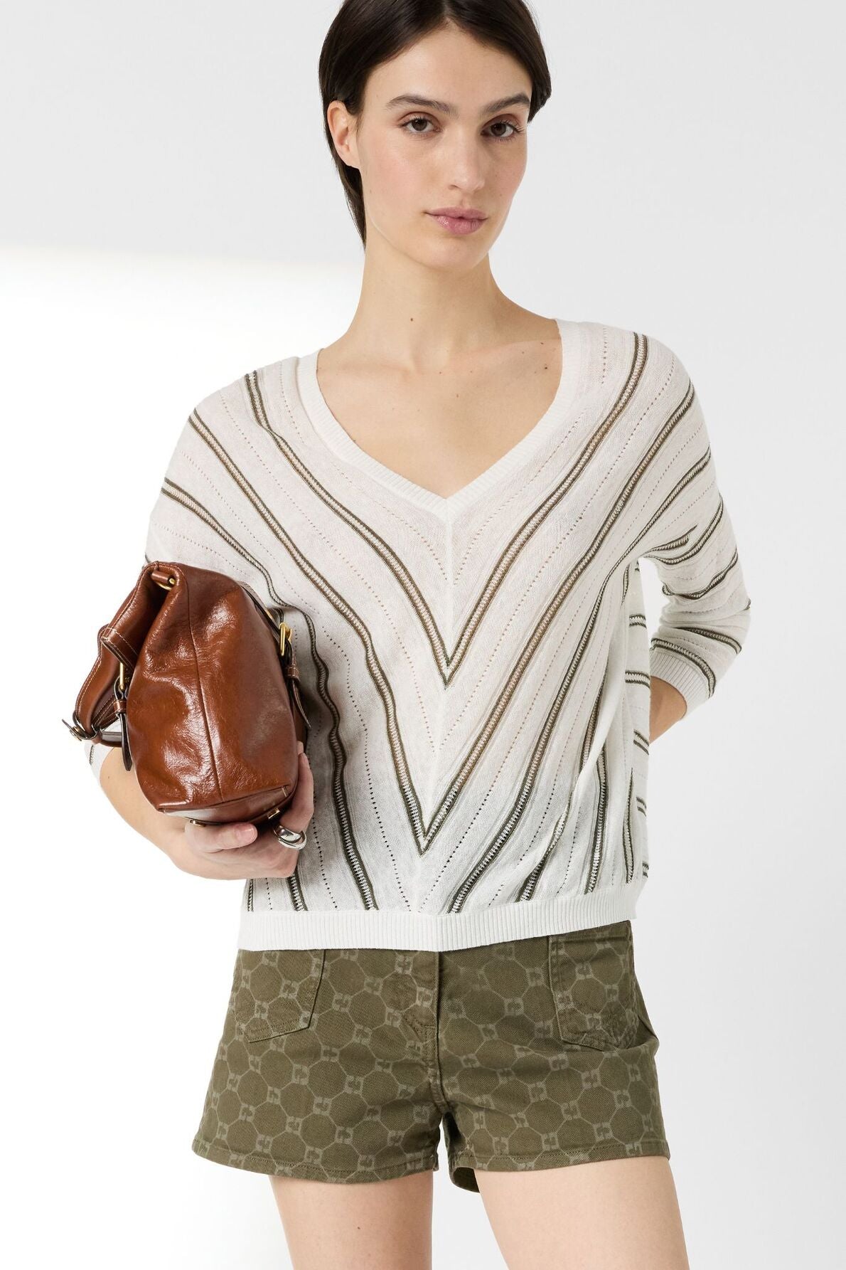 A woman wearing a pull rayé femme—a white V-neck sweater with diagonal stripes, crafted in a linen-cotton blend—pairs it with olive green patterned shorts and holds a brown leather bag. She stands against a plain white background.