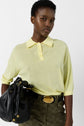 A person wearing a light yellow short-sleeved polo shirt, patterned dark green pants with a gold-buckled belt, and carrying a black handbag. They have short hair and a neutral expression against a plain background.
