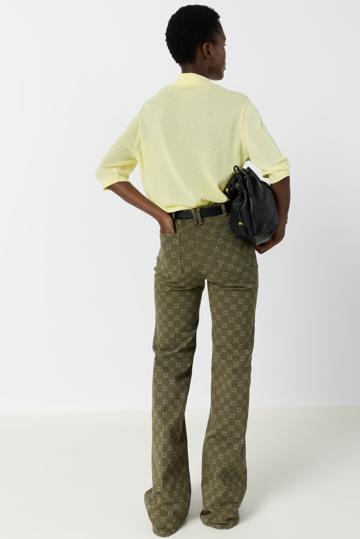 Person standing with back to camera, wearing a light yellow long-sleeve shirt, olive green patterned pants, and holding a black bag under one arm. The setting is a plain white background.