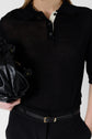 A person wearing a sheer black collared shirt, black pants with a slim black belt, and holding a shiny black handbag. Only the torso and part of one arm are visible.