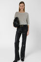 Woman standing against a white background, wearing a pull Lysia with rayures verticales in white and black, paired with black leather pants and black shoes. She holds a black handbag under her arm, her straight hair framing her neutral expression.