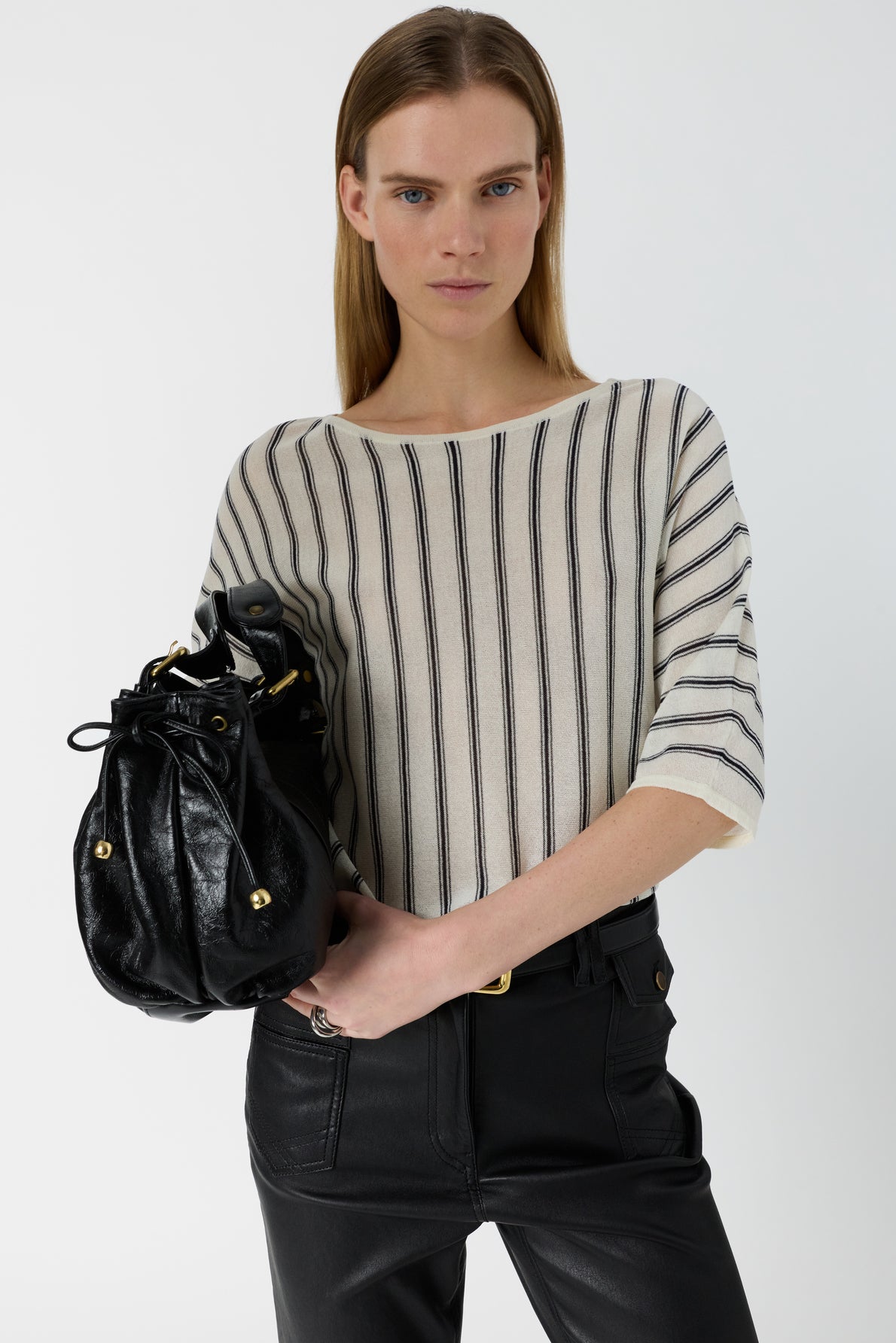 A person wearing a pull Lysia with rayures verticales and black pants holds a black leather handbag against a plain white background.