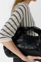 A person wearing a cream and black striped shirt with rayures verticales holds a large black leather handbag under their arm; the image is cropped to show only the torso and arm.