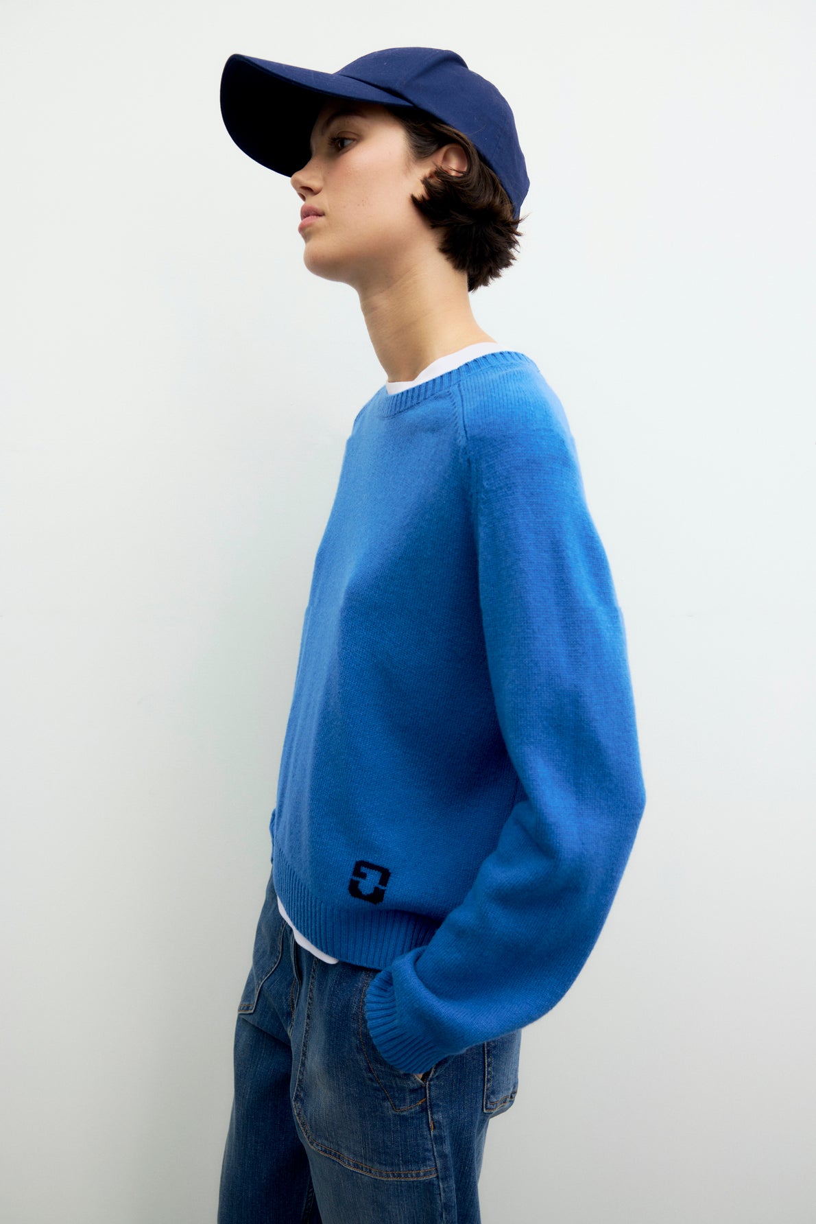 A person wearing an Esprit pull laine à col rond in blue, paired with blue jeans and a navy blue cap, stands against a plain light-colored background, looking to the side.