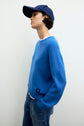 A person wearing an Esprit pull laine à col rond in blue, paired with blue jeans and a navy blue cap, stands against a plain light-colored background, looking to the side.