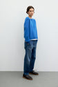 A person stands against a plain wall, wearing a loose pull laine bleu col rond layered over a white shirt, relaxed-fit blue jeans from Esprit, and dark brown lace-up shoes. Their hair is short and parted in the middle.