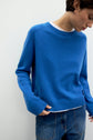 Person wearing a bright blue pull laine Esprit à col rond over a white shirt and blue jeans, standing against a plain light background, looking down with one hand in their pocket.