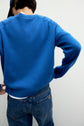 A person facing away, wearing a bright blue pull laine col rond sweater from Esprit, paired with blue jeans and a dark cap, with one arm bent resting on their hip against a plain light background.
