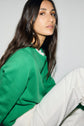 A woman with long brown hair wearing an Esprit pull laine vert à col rond and white pants sits against a plain light background, looking slightly past the camera with a calm expression.