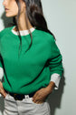 A woman with long dark hair wears an Esprit pull laine vert à col rond over a white shirt and white pants, standing against a plain light background with her hands in her pockets.