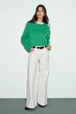 Woman standing against a plain background, wearing an Esprit pull laine vert à col rond, white wide-leg pants, and black shoes, with hands in her pockets and long dark hair loose over her shoulders.