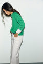 A person with long dark hair, wearing a pull laine vert Esprit à col rond over a white shirt and white pants, stands sideways with one hand in their pocket against a plain light background.