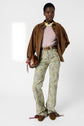 A person stands wearing a brown jacket over a light pink pull femme in cachemire with manches 3/4, floral print pants, brown belt, and brown shoes, holding a brown handbag with tassel details against a plain light background.