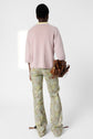 A person stands facing away, wearing a light pink pull femme à manches 3/4 en cachemire, loose floral print pants, and holding a brown handbag. The background is plain white.