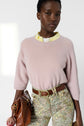 A person wearing a pale pink pull femme cachemire with manches 3/4 over a yellow collared shirt, floral pants, and a brown belt holds a brown leather handbag against a plain white background.