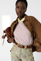 A person wearing a brown suede jacket, a cachemire pull femme with manches 3/4 in light pink, a yellow collared shirt, floral pants, and a brown belt holds a brown purse against a plain background.