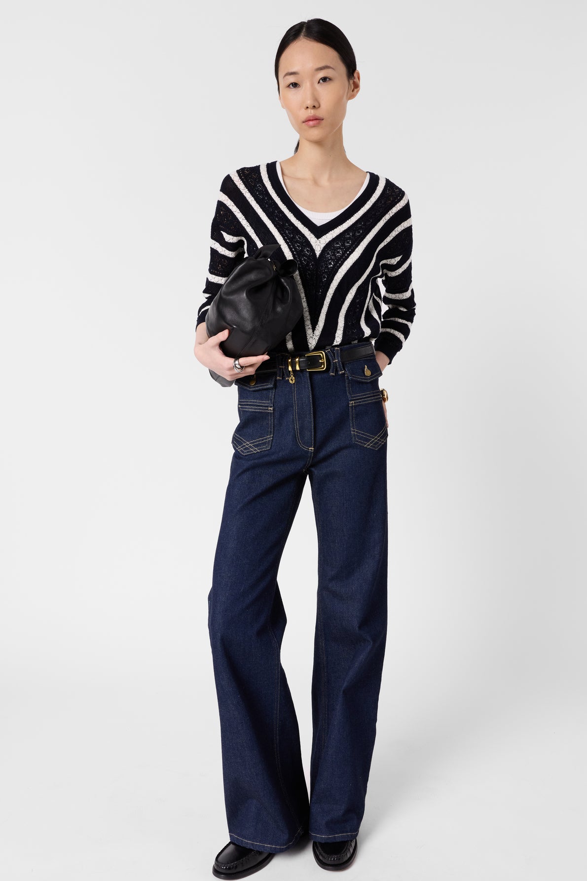 A person stands against a white background wearing a black and white pull rayé in maille ajourée, high-waisted dark blue jeans, black shoes, and holding a black handbag.