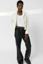 A person stands against a plain background, wearing a white textured blazer, white top, black leather pants, a black belt with a gold buckle, and a chunky gold chain necklace.
