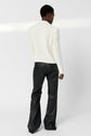 Person standing, facing away. They wear a textured white blazer and black faux leather flared pants. The background is plain white, and lighting creates a faint horizontal shadow across the wall.