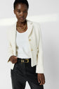 A woman wearing a white textured blazer, white ribbed top, black belted pants, and a chunky gold necklace stands against a plain light background, looking confidently at the camera.