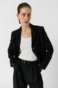 Person wearing a white ribbed tank top, a chic black plaid blazer (veste noire carreaux), and high-waisted black trousers, standing with hands in pockets against a plain white background.