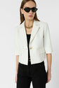 Woman wearing black sunglasses, a chunky gold necklace, a white veste tailleur courte with elbow-length sleeves featuring a subtle motif jacquard, and high-waisted black pants, standing against a plain white background.