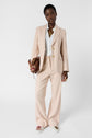 Person wearing a light beige veste nude suit with a white shirt, holding a brown handbag, and wearing white sneakers with embellishments, standing against a plain white background.