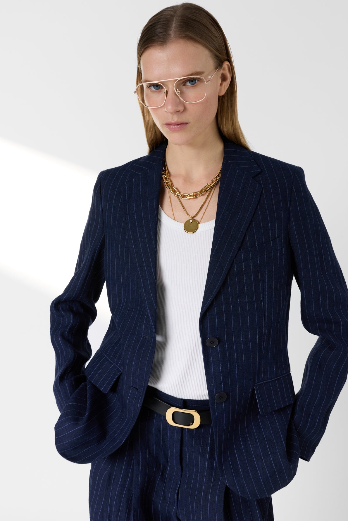 A person with long blonde hair wears glasses, a veste bleu marine à rayures, a white top, gold necklaces, and a black belt with a gold buckle, standing against a plain light background.