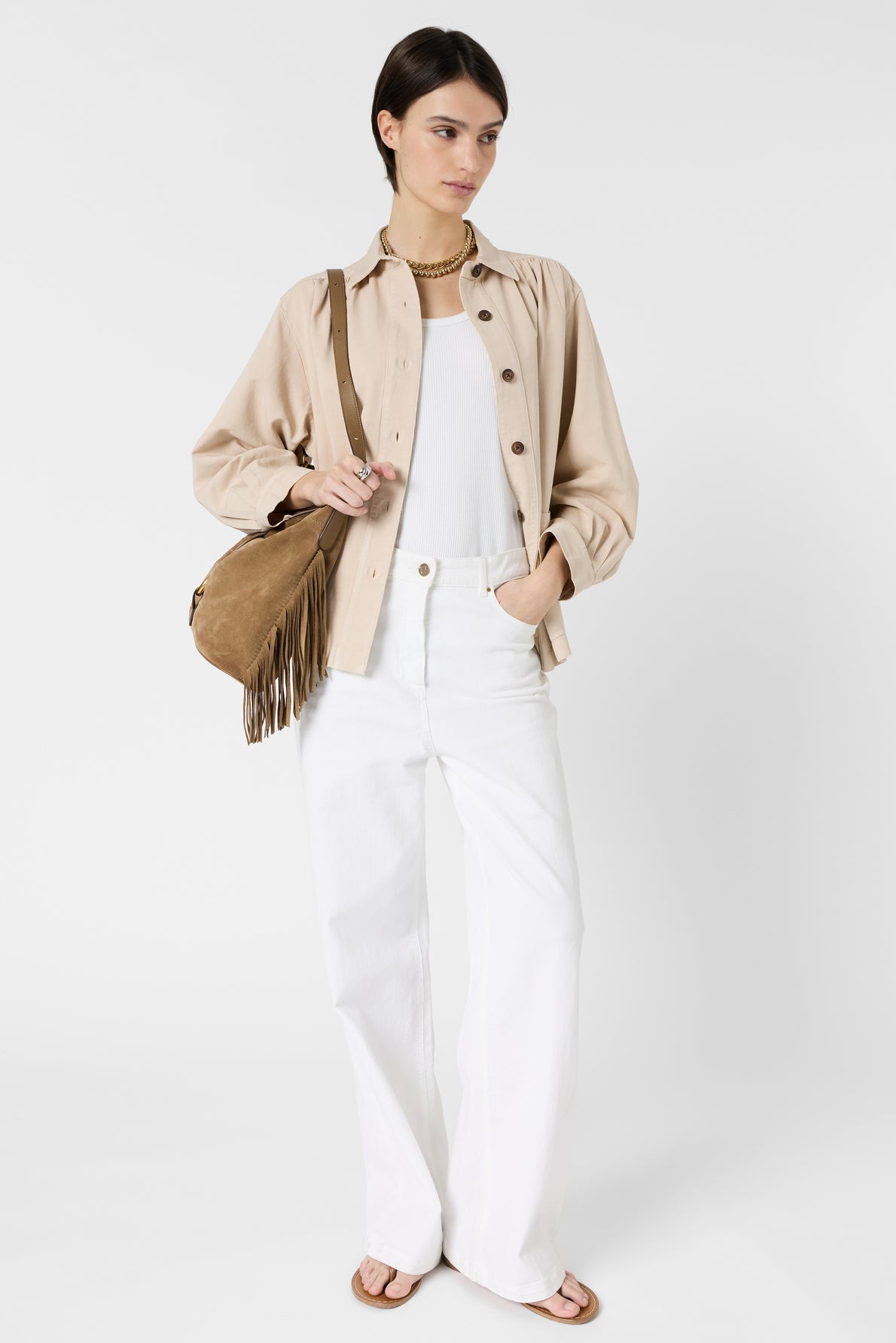 Woman standing against a white background wearing a beige surchemise lin coton over a white top, white wide-leg pants, tan sandals, and carrying a tan fringed shoulder bag. She has short dark hair and a neutral expression, creating an effortless look safari chic.