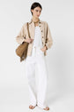 Woman standing against a white background wearing a beige surchemise lin coton over a white top, white wide-leg pants, tan sandals, and carrying a tan fringed shoulder bag. She has short dark hair and a neutral expression, creating an effortless look safari chic.