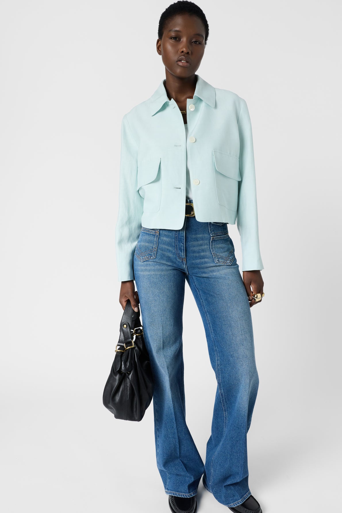 A person stands against a white background wearing a veste à col chemise in light blue, paired with a dark top, blue flared jeans, and black shoes, holding a large black handbag.