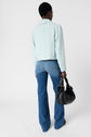 Person standing with back facing the camera, wearing a veste à col chemise light blue jacket, blue flared jeans, and holding a black handbag in the left hand. Background is plain white.