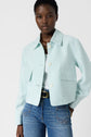 Woman wearing a veste vert pastel à col chemise over a white top and blue jeans, accessorized with a gold necklace and black belt, standing against a plain light background.