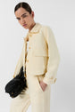 Person in a light yellow, veste jaune pastel suit jacket and pants poses with one hand in a pocket, holding a black woven bag. The background is plain white.