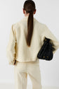 Person with long brown hair in a low ponytail, wearing a cream-colored suit and a veste courte, is shown from the back. They hold a black textured handbag under their left arm. The background is plain white.