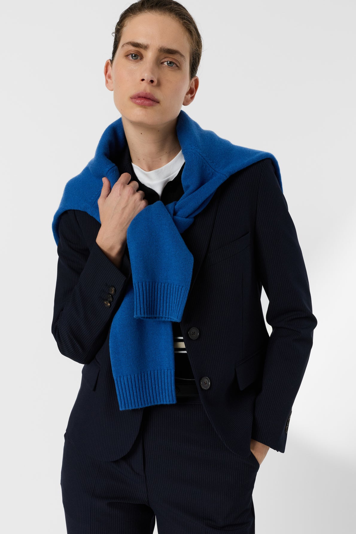 Person in a pinstripe suit with a blue sweater draped over their shoulders, holding the sweater with one hand and looking confidently at the camera against a plain background.